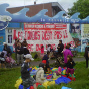 A community protest in front of a condo development sales office, with families and children gathered outdoors holding a large red banner in French.