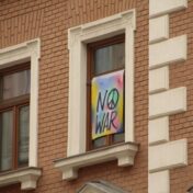 A tie dye flag saying "No War" with a peace sign hanging in a window of a brick building.