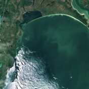 Satellite image of Lake Winnipeg's southern basin, showing the lake's teal-green water surrounded by flat agricultural land and boreal terrain in Manitoba, Canada. Partial ice cover is visible in the lower portion of the lake.