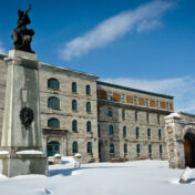 The Prison des Patriotes (Au Pied-du-Courant) in Montreal in winter, with deep snow in the foreground, a stone monument dedicated to the Patriotes in the left foreground, and a blue sky overhead.
