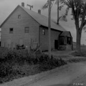 Black and white archival photograph of a wood-shingled barn or farmhouse with a metal roof, two chimneys, and a weathervane, seen from the roadside. A wooden fence and overgrown brush line a dirt road in front of the building, with tall trees to the right. The image is labeled 'T.S. 9131' in the bottom corner.