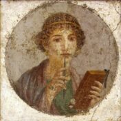A woman dressed in clothing styled after first-century Rome (roughly 55–79 AD) stands holding a set of wax tablets in one hand and a stylus in the other, as if pausing mid-writing.