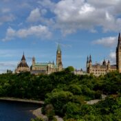 A view of Canada's Parliament Hill in Ottawa, seen across the Ottawa River from a distance, with green, tree-covered slopes in the foreground. The Gothic Revival sandstone buildings include the Library of Parliament's polygonal tower on the left, the Peace Tower with its clock and Canadian flag at center, and the East Block's spires on the right, all set against a blue sky with scattered white clouds.