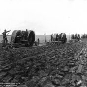 A black-and-white photograph showing a line of steam traction engines plowing a prairie field near Saskatoon, Saskatchewan, circa early 1900s. Several operators tend the large wheeled machines as they cut furrows across flat, open farmland. Photographed by B.P. Skewis of Saskatoon; copyrighted in Canada and USA