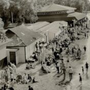 Black-and-white archival photograph, taken from an elevated vantage point, of a crowded lakeside beach lined with wooden bathhouses and a large pavilion with a peaked roof. Dozens of people in early 20th-century swimwear and street clothes stand, sit, and walk along the sandy shore, with several wading or swimming near a diving platform and slide in the water on the right. Tall lampposts and leafy trees frame the scene.