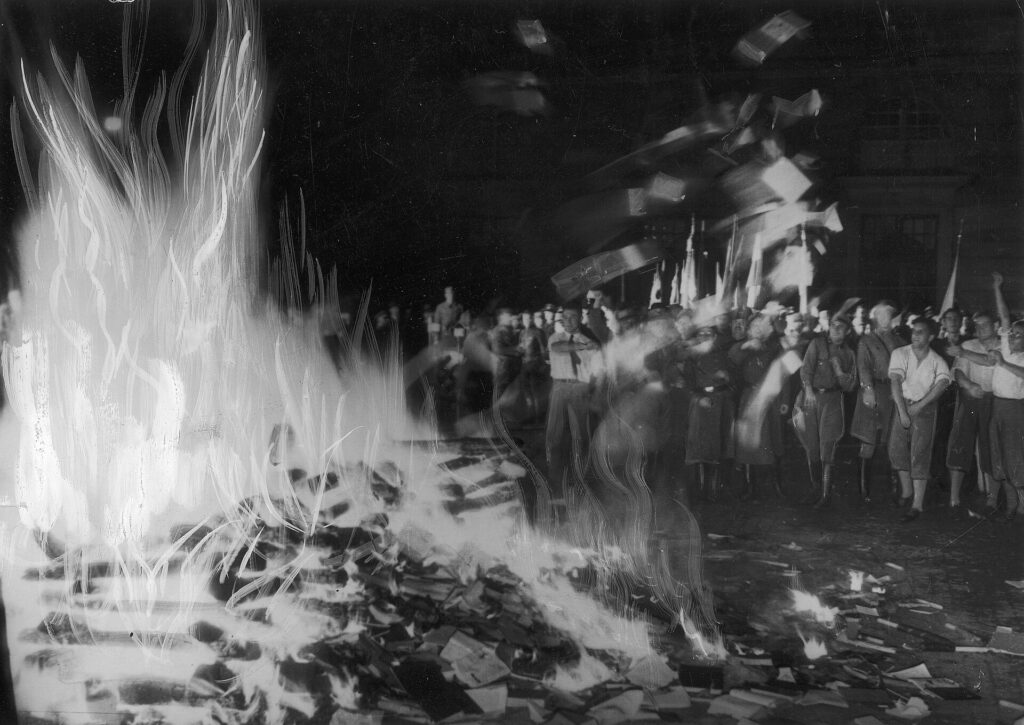 Black and white photo of a crowd of people, some in military uniforms, throwing books into a huge bonfire.