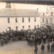 Sepia photo of a large group of people in dark suits and hats facing away from the camera, toward a large building. There is a large stone building under construction on the right side of the image.