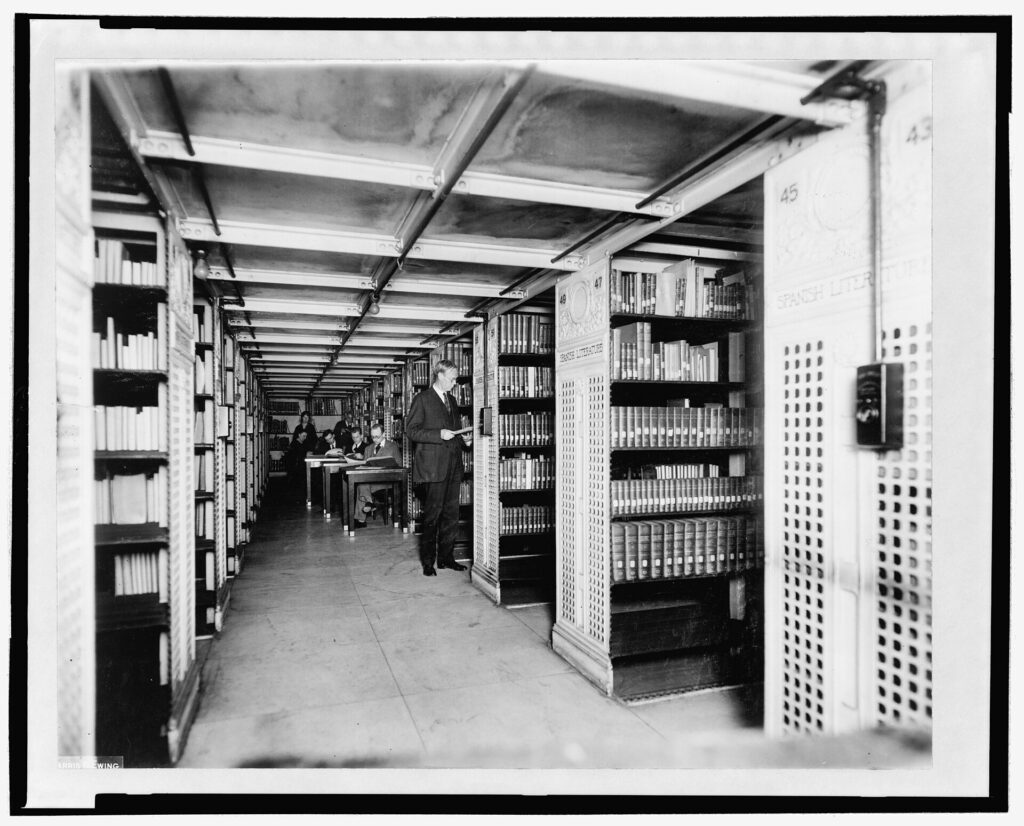 Black and white photograph of a library hallway lined with bookshelves. There is one person standing at a shelf and several people in the background are seated at desks, reading.