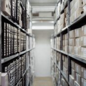 Colour photo of the lane between two sets of metal shelves in an archive. The shelves are filled with labeled boxes.
